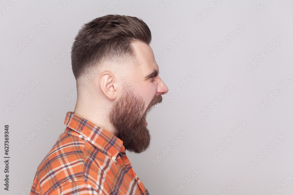Side view portrait of serious strict angry bearded man standing looking ahead, screaming with aggression, wearing orange checkered shirt. Indoor studio shot isolated on gray background. Side view portrait of serious strict angry bearded man standing looking ahead, screaming with aggression, wearing orange checkered shirt. Indoor studio shot isolated on gray background.
