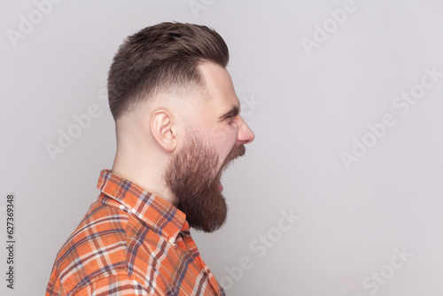 Photos Side view portrait of serious strict angry bearded man standing looking ahead, screaming with aggression, wearing orange checkered shirt