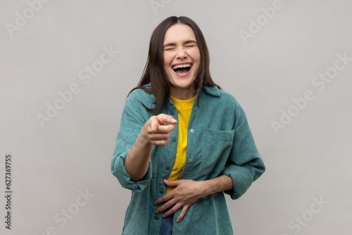 Joyful woman laughing, holding stomach, pointing to camera, taunting you, can't stop hysterical laughter, wearing casual style jacket. Indoor studio shot isolated on gray background.