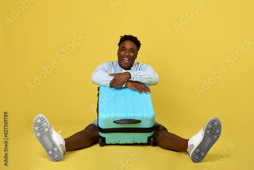 On a yellow background, a handsome man in a long-sleeved shirt sat and folded his hands over a large travel suitcase. Cheerful dark-skinned guy posing with a suitcase on wheels in a yellow studio