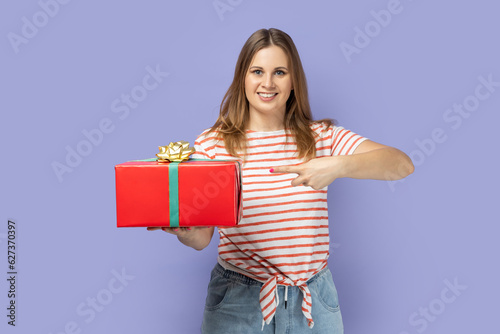 Portrait of pretty good looking attractive blond woman wearing striped T-shirt holding and pointing finger at red present box, smiling to camera. Indoor studio shot isolated on purple background.