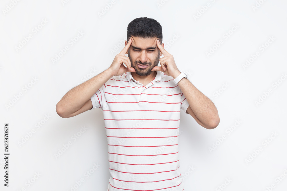 Portrait of sick unhealthy ill bearded man wearing striped t-shirt standing suffering headache, massaging his temples, frowning face. Indoor studio shot isolated on gray background. Portrait of sick unhealthy ill bearded man wearing striped t-shirt standing suffering headache, massaging his temples, frowning face. Indoor studio shot isolated on gray background.