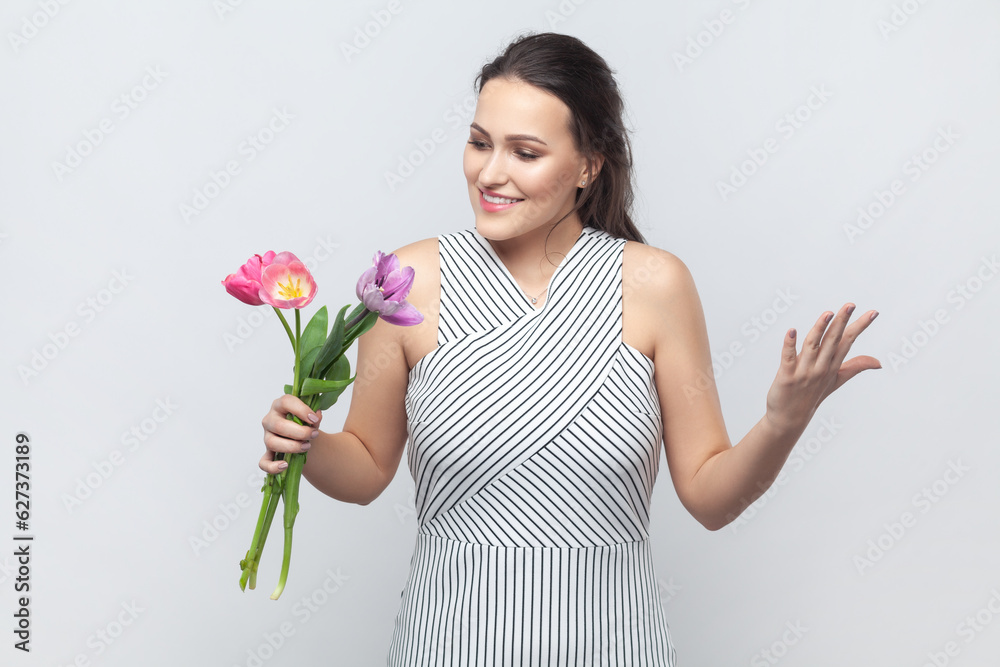 Portrait of amazed happy smiling brunette woman gets bouquet of spring flowers, holding flowers, being glad, pleasant surprise, wearing striped dress. Indoor studio shot isolated on gray background. Portrait of amazed happy smiling brunette woman gets bouquet of spring flowers, holding flowers, being glad, pleasant surprise, wearing striped dress. Indoor studio shot isolated on gray background.