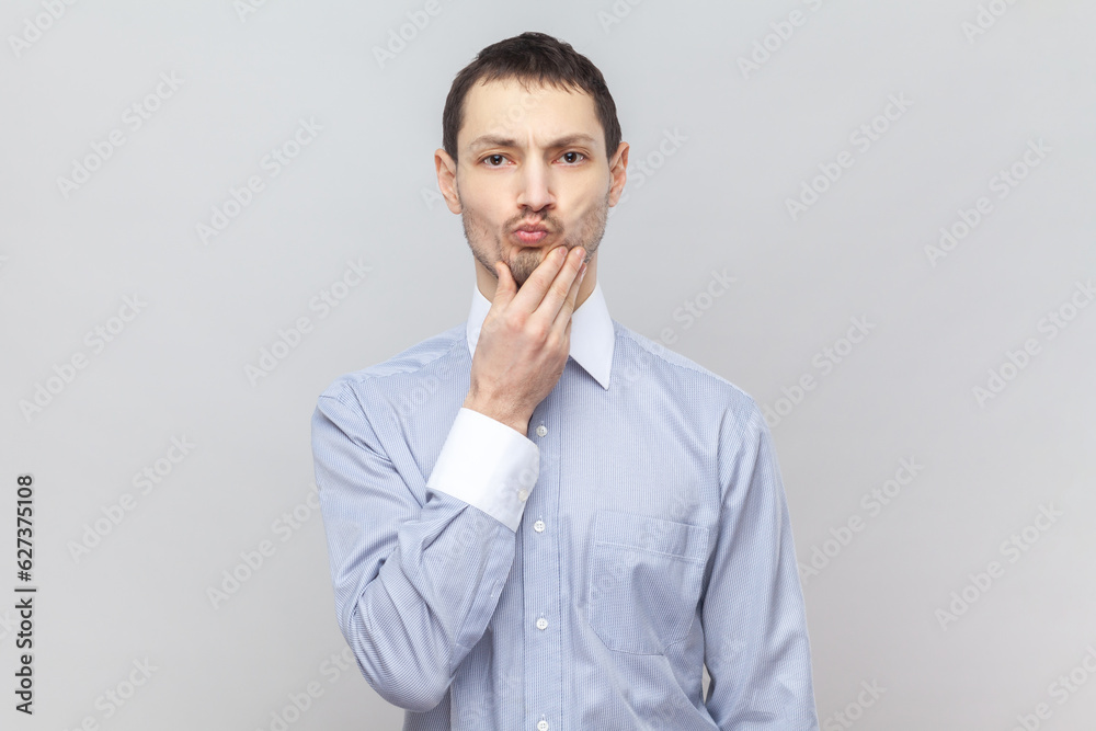 Portrait of serious pensive man keeps hand on his chin, looking at camera, thinking about something important, planning, wearing light blue shirt. Indoor studio shot isolated on gray background. Portrait of serious pensive man keeps hand on his chin, looking at camera, thinking about something important, planning, wearing light blue shirt. Indoor studio shot isolated on gray background.