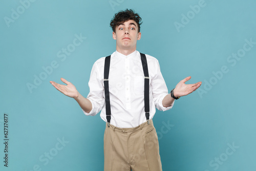 Portrait of puzzled confused man wearing white shirt and suspender, shrugging shoulders, looking away, raised his arms, has uncertain expression. Indoor studio shot isolated on blue background.
