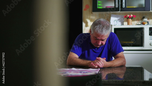 Preoccupied senior man at kitchen table ruminating problems, candid domestic scene of elderly person going through difficulties and loneliness