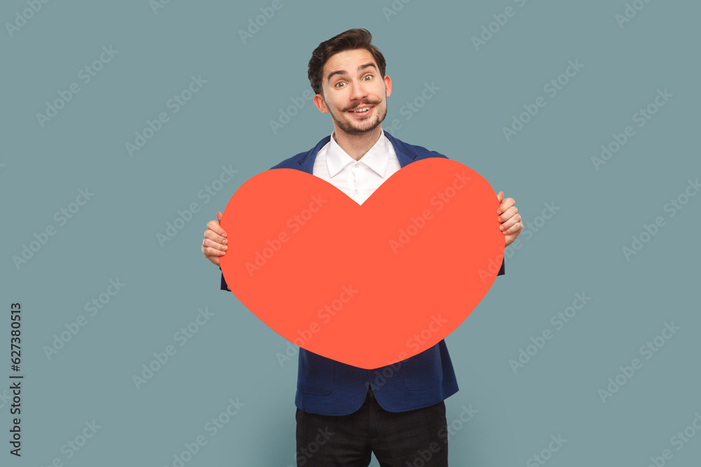 Portrait of positive excited amazed man with mustache standing holding big red heart, expressing romantic feelings, wearing white shirt and jacket. Indoor studio shot isolated on light blue background Portrait of positive excited amazed man with mustache standing holding big red heart, expressing romantic feelings, wearing white shirt and jacket. Indoor studio shot isolated on light blue background