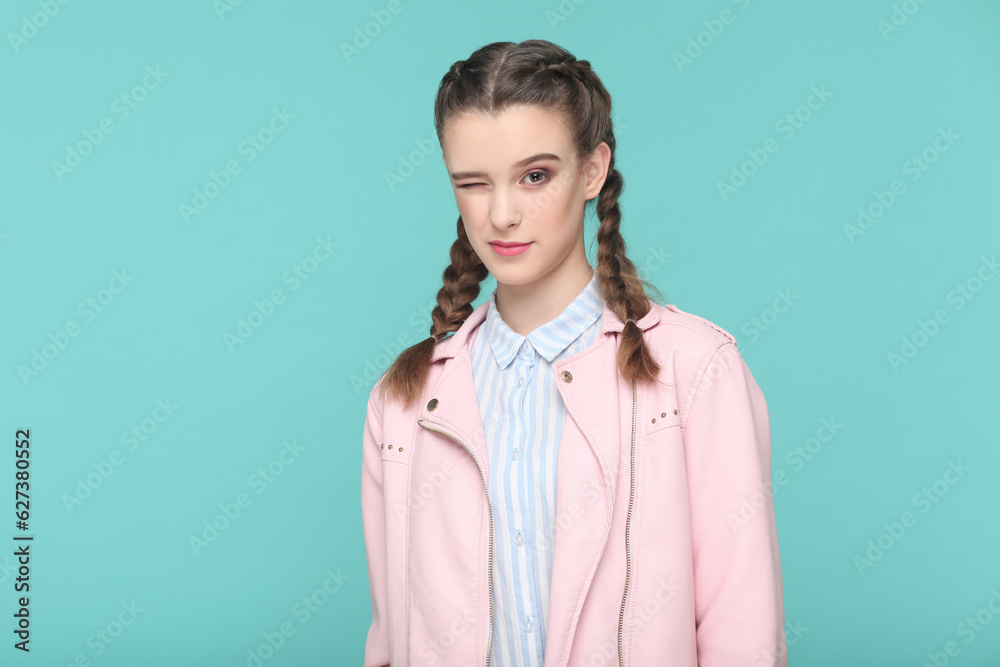 Portrait of playful cheerful teenager girl with braids wearing pink jacket standing looking at camera, winking, flirting with boyfriend. Indoor studio shot isolated on green background. Portrait of playful cheerful teenager girl with braids wearing pink jacket standing looking at camera, winking, flirting with boyfriend. Indoor studio shot isolated on green background.