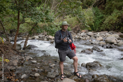 Man standing on the rocky shore of a white water river photographing the view in the Iao Valley in Central Maui; Maui, Hawaii, United States of America