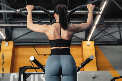Rear view of sporty woman workout pulling up on bar in fitness gym. Lady exercising pulls herself in modern sports center, working out. Healthy lifestyle and sport training concept. Copy ad text space