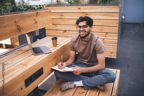 Smiling man resting on wooden bench and writing in notepad in cafe