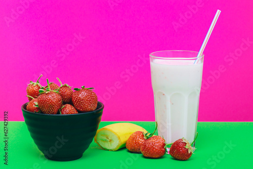 Black bowl with strawberries, glass of cold milkshake, banana on green and pink background.