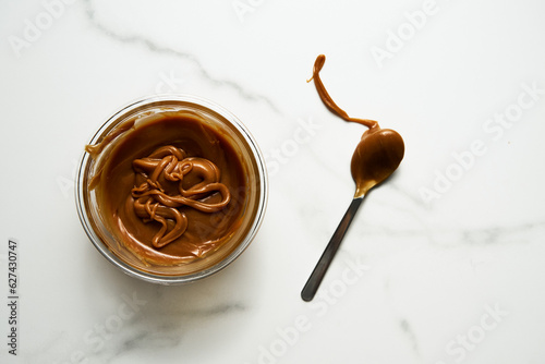 Salted Caramel sauce in glass bowl on white marble background. Next to it lies a spoon soiled in caramel. Butter, sugar with cream and salt. Top view food photo.