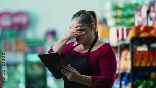 Stressed female entrepreneur owner of small business struggling with hard times holding tablet and staring at the bottom line of the grocery store