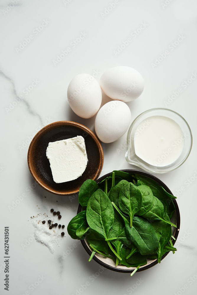 Close up of iingredients for healthy spinach and feta omelette. Eggs with heavy cream, Fresh baby spinach leaves in the black ceramic bowl, white marble background. Top view food photo Close up of iingredients for healthy spinach and feta omelette. Eggs with heavy cream, Fresh baby spinach leaves in the black ceramic bowl, white marble background. Top view food photo