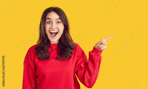 Portrait of young excited woman with opened mouth in red sweatshirt pointing on copy space on yellow background looking at camera. Sincere human emotions concept. Banner for advertisement, marketing.