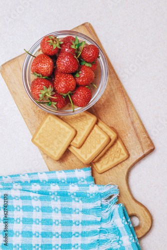 Fresh ripe strawberry in a glass bowl and cookies at wooden board on a light table. Heap of strawberries. Top view.