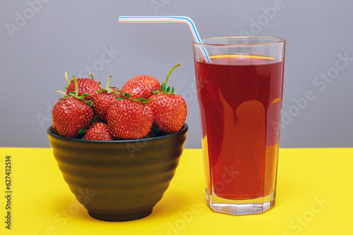 Fresh strawberry and juice. Black bowl with strawberries and glass of red juice on yellow and gray background.