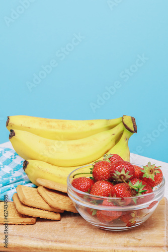Strawberry, bananas and cookies lies at wooden board on light table with blue background.