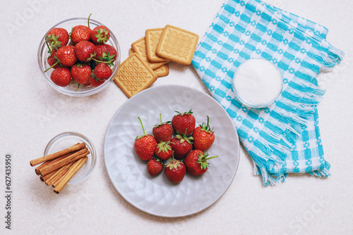 Heap of freshly harvested strawberries on a light table. Strawberries served in a ceramic plate and glass bowl with bowl of sugar, cinnamon and cookies. Top view.
