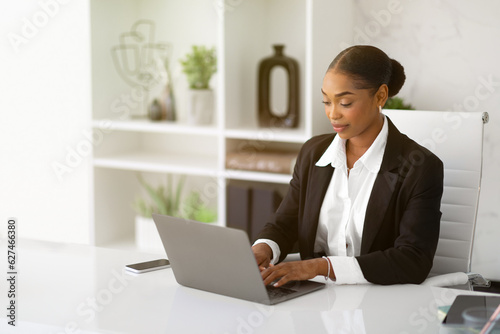 Beautiful black businesswoman in formal wear working with laptop computer at desk in office interior, copy space