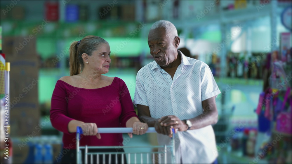 Diverse customers conversing inside grocery store with shopping cart. African American senior speaking with a caucasian woman inside business shop