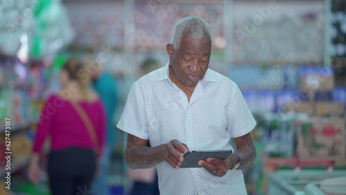 One black senior Brazilian manager of grocery store holding tablet device posing for camera inside small business. Older African American person using modern technology © Marco