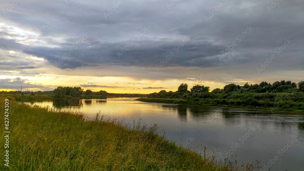 The sun sinks below the horizon on a summer evening. The colorful cloudy sky and trees are reflected in the river water. The high bank of the river is overgrown with grass. A forest grows