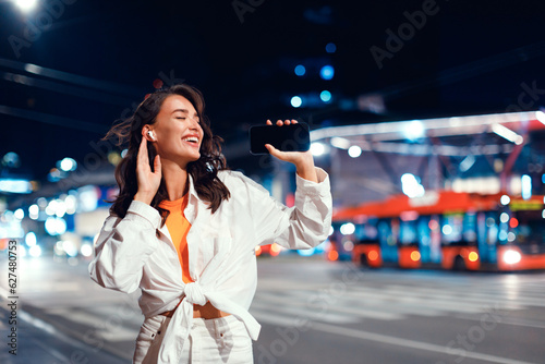Smiling caucasian woman using mobile phone and earphones while walking in the city at night, free space