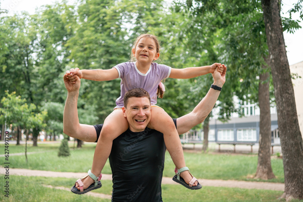 Child girl piggybacks on father, riding on father's shoulder in park. Happy family bonding time, fatherhood concept 