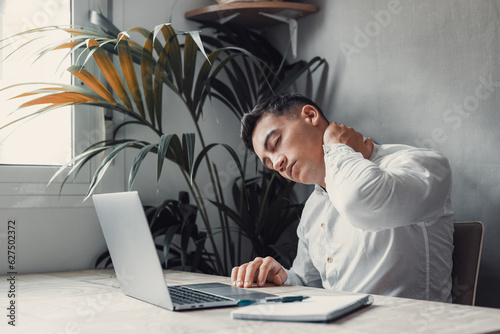 Exhausted young Caucasian male worker sit at desk massage neck suffer from strain spasm muscles. Tired unwell man overwhelmed with computer work sedentary lifestyle struggle with back pain or ache..