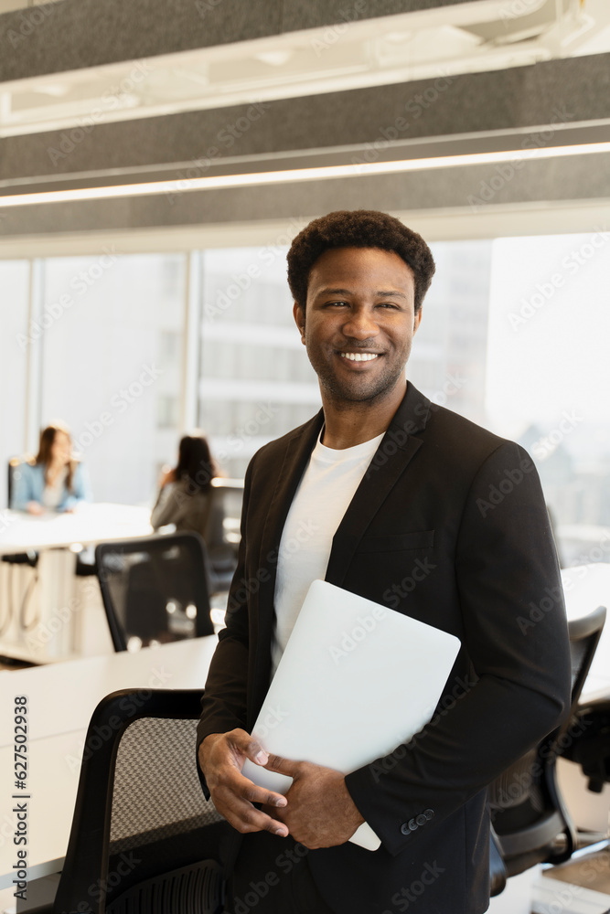 Portrait of confident smiling African American man, manager holding laptop computer, planning project, looking away standing in modern office. Successful business, career concept 	