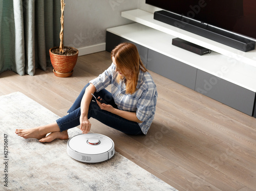 The girl sits on the floor and controls the robot vacuum cleaner using a smartphone, smart home.