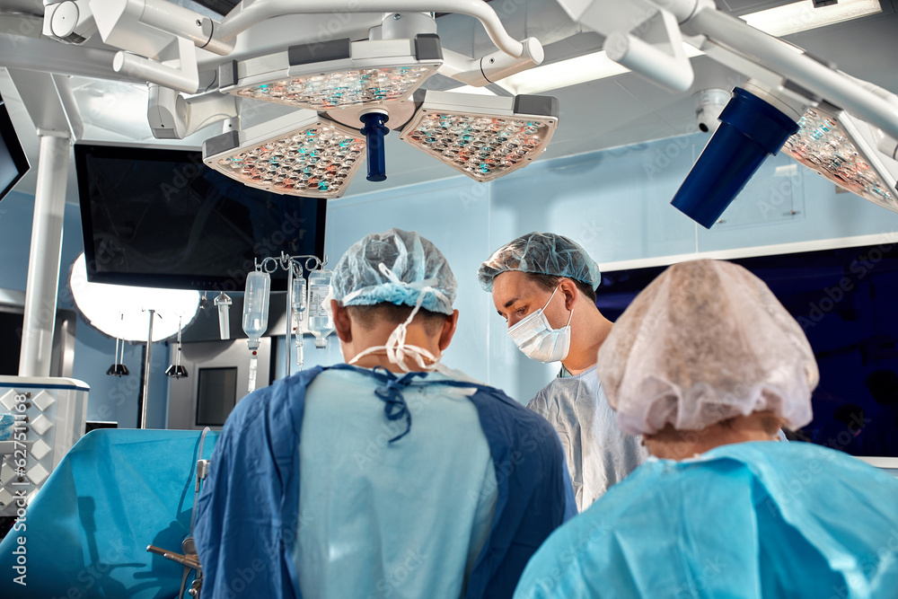 Surgeons team during complex surgical operation in a bright sterile operating room. Doctors leaned over patient using modern surgical instruments and clamps. Precision medicine, saving patient's life. Surgeons team during complex surgical operation in a bright sterile operating room. Doctors leaned over patient using modern surgical instruments and clamps. Precision medicine, saving patient's life.