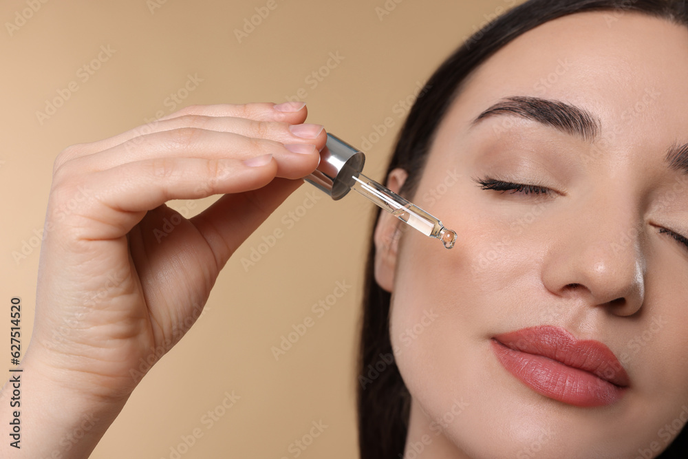 Young woman applying essential oil onto face on beige background, closeup