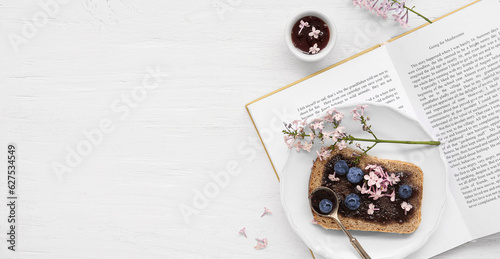 Book, plate with sweet blueberry jam toast and flowers on white wooden background with space for text