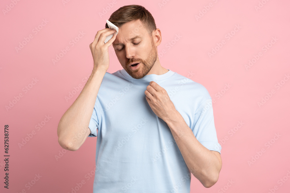 Overheated sweaty man suffering from heat stroke from high weather temperature using paper napkin for forehead. Dehydrated guy feeling unhealthy need air conditioner isolated on pink studio background Overheated sweaty man suffering from heat stroke from high weather temperature using paper napkin for forehead. Dehydrated guy feeling unhealthy need air conditioner isolated on pink studio background