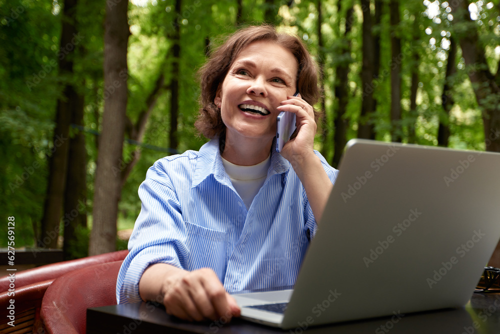 Adorable mature woman with laptop and smartphone sitting at the table in outdoor cafe in the park. Middle-aged smiling brunette in a blue shirt talking on cell phone. Active lifestyle for all ages. Adorable mature woman with laptop and smartphone sitting at the table in outdoor cafe in the park. Middle-aged smiling brunette in a blue shirt talking on cell phone. Active lifestyle for all ages.