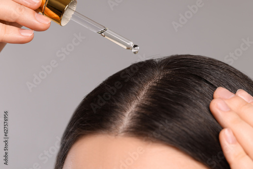 Woman applying essential oil onto hair roots on light grey background, closeup