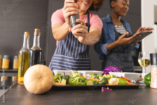 Happy diverse couple preparing meal, using tablet and seasoning vegetables in kitchen