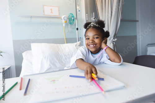 Portrait of happy african american girl patient lying on bed colouring in patient room at hospital