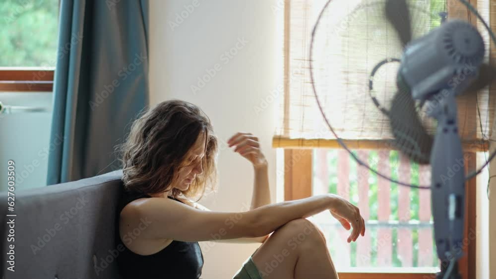 A young Caucasian woman tired from summer heat sitting on the floor in front of a working electric fan.