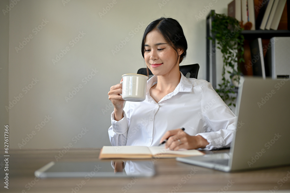 A happy Asian businesswoman enjoying her morning coffee at her desk in her office. A happy Asian businesswoman enjoying her morning coffee at her desk in her office.