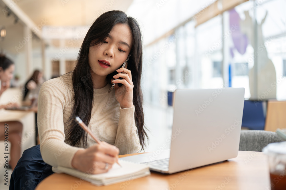 A beautiful Asian businesswoman is taking notes in her notebook and having a serious phone call