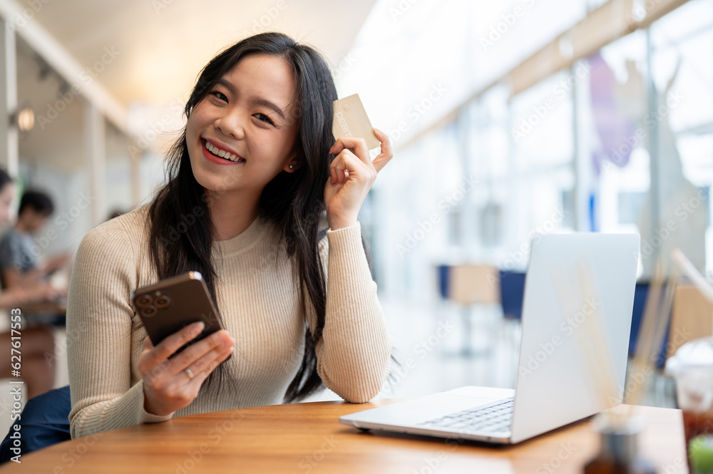 A happy Asian woman holding her smartphone and a debit card while sitting at a table in a cafe A happy Asian woman holding her smartphone and a debit card while sitting at a table in a cafe