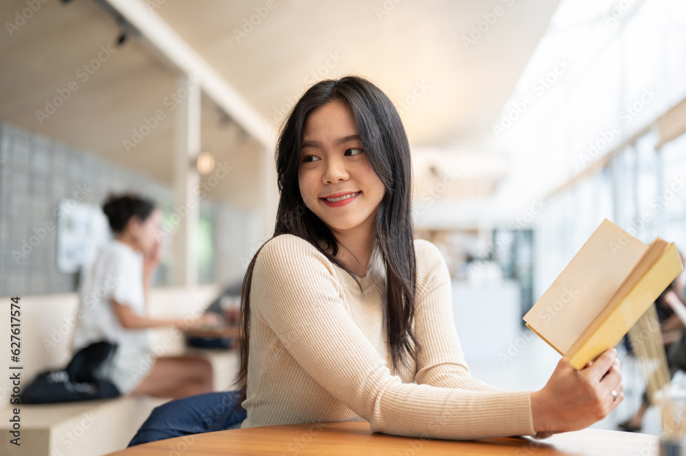 An attractive Asian woman in casual clothes poses with a book while sitting in a coffee shop.