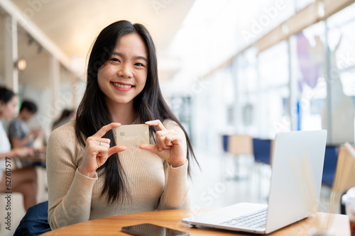 A happy Asian woman showing her debit card to the camera while sitting at a table in a coffee shop