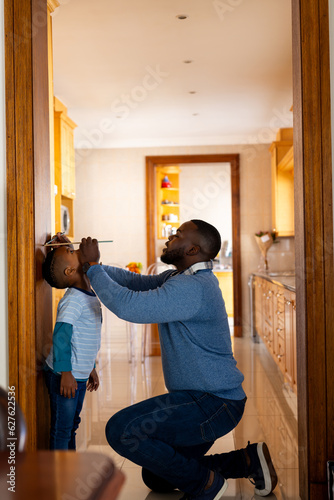 Happy african american father measuring son and marking height on door frame at home, copy space