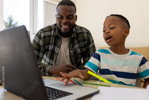 Happy african american father and son sitting table using laptop and doing schoolwork together