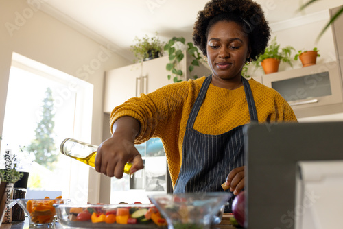 Plus size african american woman in apron pouring oil on vegetables in kitchen